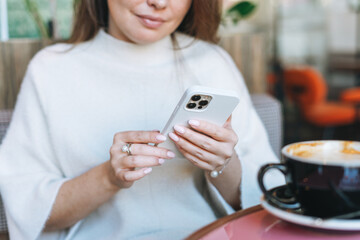 Fototapeta premium Attractive young brunette smiling woman in white casual dress with cup of coffee using mobile phone in cafe
