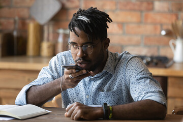 Close up serious African American man wearing glasses recording voice message, holding phone near...