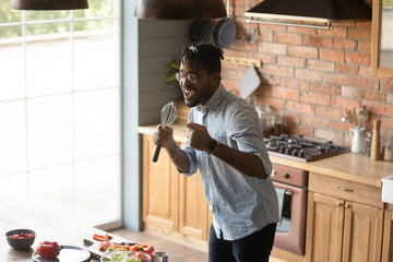 Funny African American man wearing glasses singing into whisk, dancing, having fun in kitchen,...