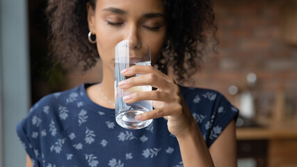 Close up satisfied African American young woman enjoying, drinking pure fresh mineral water with closed eyes, holding glass, healthy lifestyle and good daily habit concept, body and skin care - Powered by Adobe