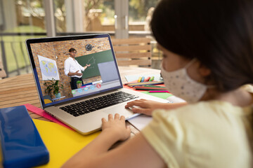 Female student wearing face mask having a video call with male teacher on laptop at school