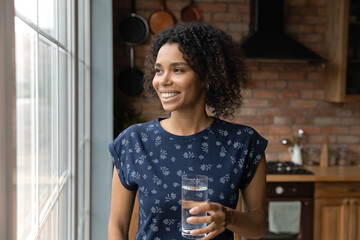 Close up dreamy smiling African American woman holding glass of pure fresh mineral water, standing...