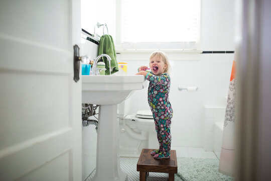 Portrait of baby girl brushing teeth while standing on stool in bathroom