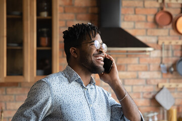 Head shot close up overjoyed African American man wearing glasses talking on phone, standing in kitchen at home, excited young male enjoying pleasant conversation, chatting, sharing news with friend