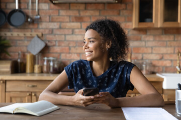 Close up smiling dreamy African American woman distracted from phone, online chatting, looking to aside, holding smartphone, waiting for call, visualizing future, thinking about new opportunities