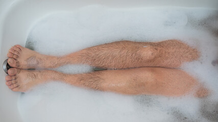 Close-up of male hairy legs in foam. A faceless man is taking a relaxing bath. Top view