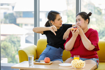 Asian fat woman feels guilty when she wants to eat a donut while her friend sitting beside her and thumb down to tell her that it is not right to eat dessert