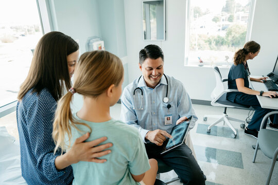 High angle view of pediatrician showing x-ray image on tablet computer to mother and daughter while female doctor working in background