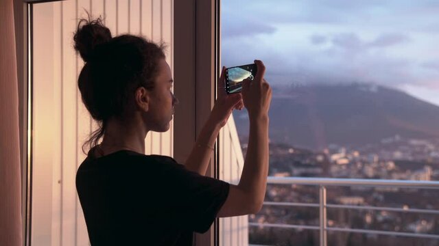 A young woman in home clothes with her hair pulled back photographs a beautiful sunset over the sea through the window of her hotel room. Beautiful sunrise over the seaside town