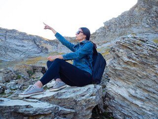 young female tourist with a backpack sits on a rock in a mountain valley against the background of peaks,