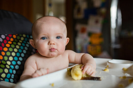 Portrait of cute shirtless baby girl with banana sitting on high chair