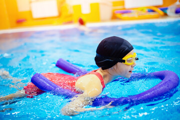 Child girl is engaged in swimming with trainer in pool, uses polyurethane stick to practice stroke