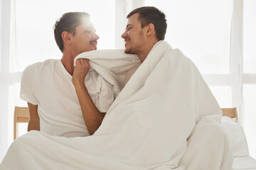 Fototapeta premium Happy young men wrapping in warm blanket and smileing at each other while resting on comfortable bed in morning in cozy bedroom