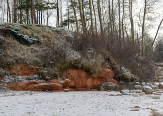 winter landscape with sandy beach, wild sea shore, red sand and clay outcrops on the sea shore, tree silhouettes on the shore, Vidzeme rocky sea shore
