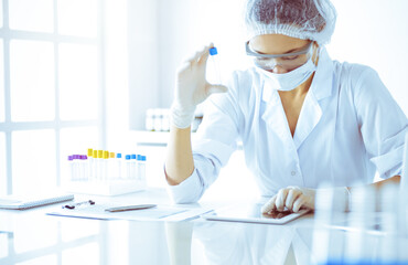 Professional female scientist in protective eyeglasses researching tube with reagents in sunny...