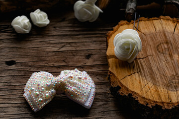 Jewelry and hair accessories. Elegant bow with holographic stones and rhinestones with a pink tint. There are cute milky roses around. Costume jewelry on an old wooden background covering