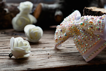 Jewelry and hair accessories. Elegant bow with holographic stones and rhinestones with a pink tint. There are cute milky roses around. Costume jewelry on an old wooden background covering