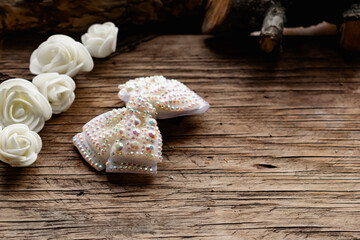 Jewelry and hair accessories. Elegant bow with holographic stones and rhinestones with a pink tint. There are cute milky roses around. Costume jewelry on an old wooden background covering