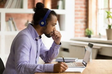 Pensive young African American man in headphones look at laptop screen thinking studying online from home. Thoughtful ethnic male student in earphones take distant course on computer learning.