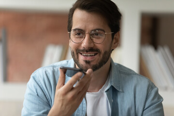 Close up of smiling young Caucasian man in glasses talk speak on smartphone on loudspeaker with client. Happy male employee record audio message, use digital voice assistant on cellphone gadget.