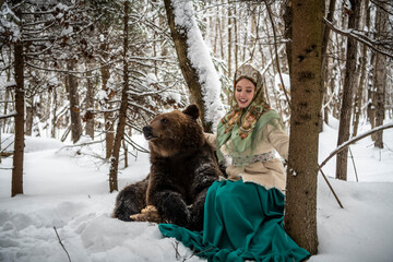 Russian beauty in folk national dress collects snowdrops in the winter forest with a brown bear