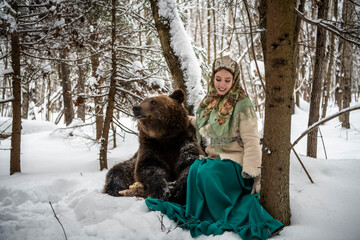 Russian beauty in folk national dress collects snowdrops in the winter forest with a brown bear