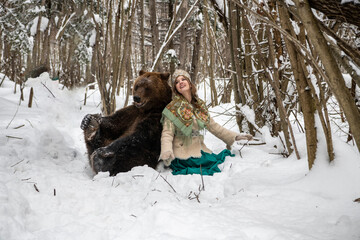 Russian beauty in folk national dress collects snowdrops in the winter forest with a brown bear