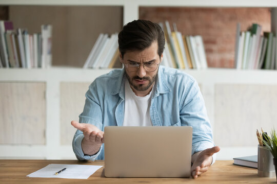 Unhappy Caucasian Male Employee Work Online On Laptop In Office Frustrated By Error Or Mistake On Gadget. Upset Mad Man Worker Look At Computer Screen Confused By Slow Internet Or Device Breakdown.