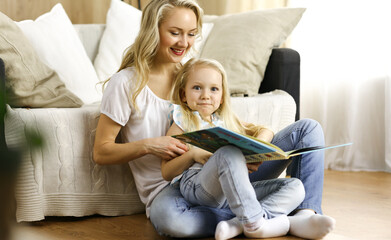 Happy family. Blonde young mother reading a book to her cute daughter while sitting at wooden floor. Motherhood concept