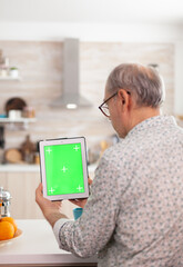 Back view of mature man holding tablet pc with chroma key in kitchen during breakfast. Authentic...