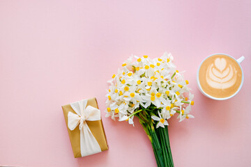 Lush bouquet of daffodils, cup of cappuccino and present in golden wrapping paper isolated on pink background. Tender minimalistic spring flowers composition. Top view, copy space, flat lay, close up.
