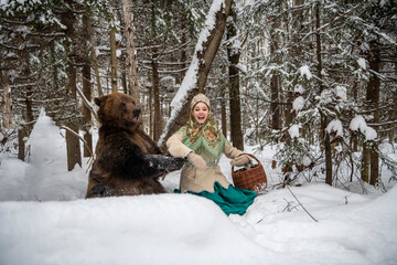 Russian beauty in folk national dress collects snowdrops in the winter forest with a brown bear