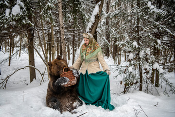 Russian beauty in folk national dress collects snowdrops in the winter forest with a brown bear