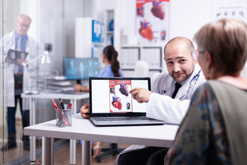 Young doctor explaining heart disease pointing on laptop sitting in hospital room talking with senior woman. Team of doctors working in background using clipboard, body scan and x-ray of sick patient