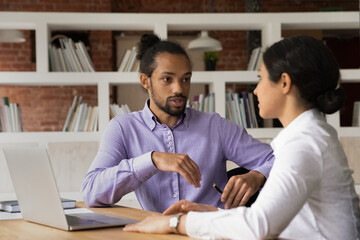 Young diverse multiracial employees sit at desk in office talk discuss company project using...