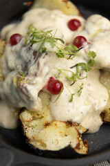 Fried potatoes in a frying pan with cream gravy and berries close-up. Potatoes baked in coals with herbs, restaurant serving of the dish.