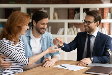 Smiling male real estate agent or broker give keys to excited young Caucasian couple buy first shared house together. Happy man realtor congratulate overjoyed spouses client with new home rental.