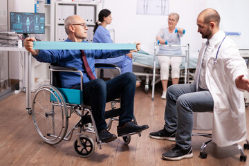 Pensioner in wheelchair doing treatment with elastic band with help from medical staff. Elderly...