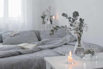 burning candles and eucalyptus in glass vase in white bedroom