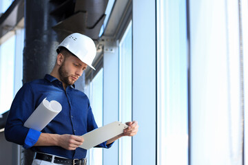 Confident young business man in hardhat holding blueprint and looking at documents while standing indoors.