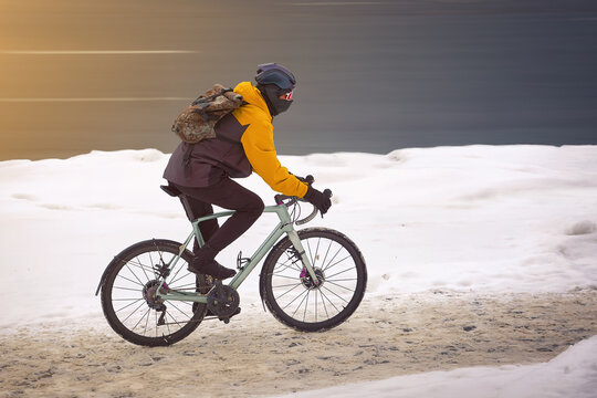 Man rides bicycle on snowy walkway in winter day. Man in warm clothes wearing protective helmet riding on snowy slippery road on bicycle. Dangerous riding bike on uncleaned path from snow