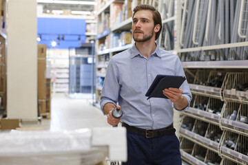 Young man shopping or working in a hardware warehouse standing checking supplies on his tablet.