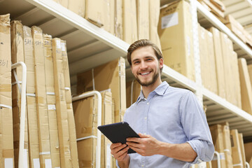 Young man shopping or working in a hardware warehouse standing checking supplies on his tablet. © ty