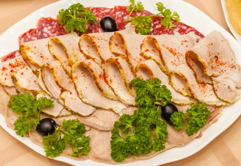 Fototapeta na wymiar Sliced boiled meat, sausage, black olives and sprigs of parsley close-up on a white plate on a tablecloth background