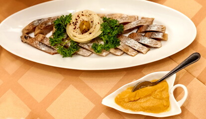 Sliced salted fish herring decorated with a sprig of parsley, onion rings and green olive on an oval white ceramic plate and a plate with mustard and spoon close-up on the tablecloth background