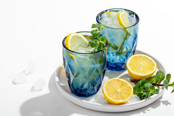 Summer lemonade with mint and ice in blue glasses on a white table on a plate, next to lemons, ice and mint, selective focus on mint, copy space
