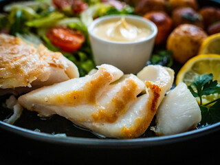 Fish dish - fried cod fillet with potatoes and vegetable salad on black wooden table