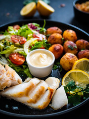 Fish dish - fried cod fillet with potatoes and vegetable salad on black wooden table