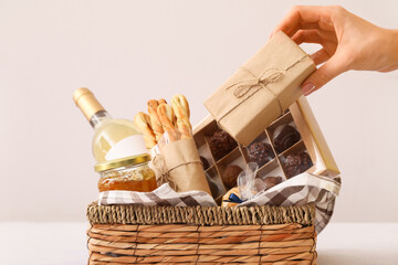 Woman putting gift in basket with products on light background