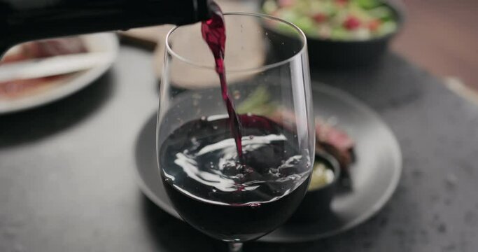 Slow motion closeup pour red wine in wineglass with steak on background